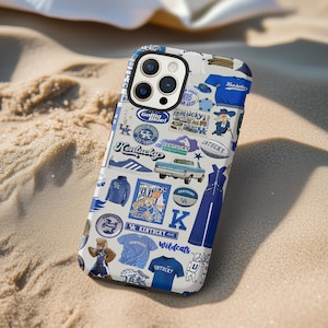 May include: A white and blue phone case featuring various Kentucky Wildcats graphics and text. The design includes logos, text that says "Kentucky", and images of a truck, basketball player, and other related items. The case is on a sandy surface.