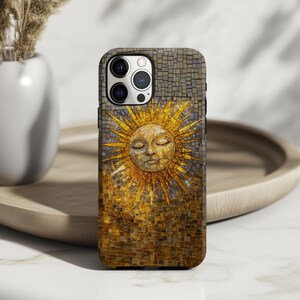 May include: A phone case featuring a mosaic sun design with a smiling face and radiant rays. The case has a textured, multi-colored appearance with shades of gold, brown, and gray. The phone case is on a wooden tray.