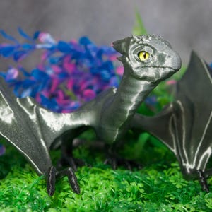 May include: A detailed, gray dragon figurine with yellow eyes and outstretched wings. The dragon is posed on a bed of green faux grass, with blue and purple artificial plants in the background. The dragon's wings have white accents.