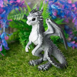 May include: A detailed figurine of a dragon with gray and white scales, black claws, and large wings. The dragon is posed on a bed of green moss, with purple and blue artificial flowers in the background. Fantasy decor.
