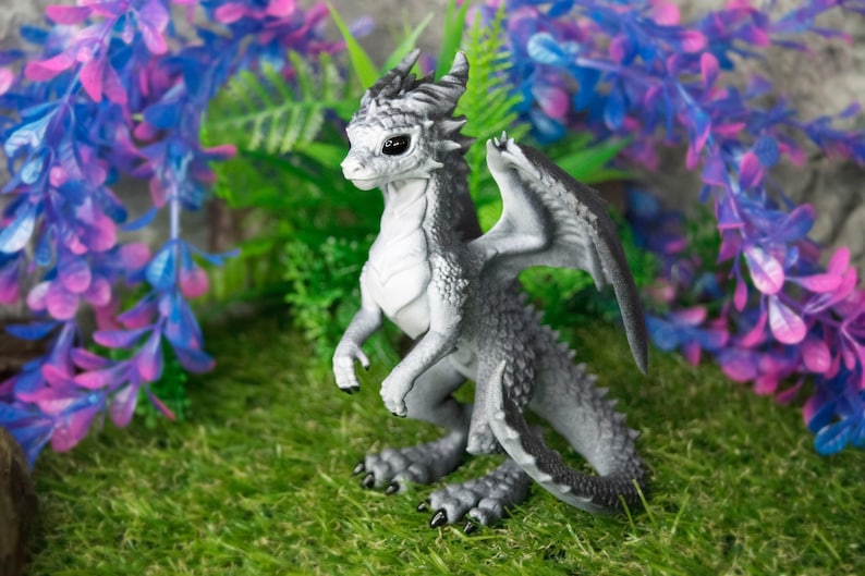 May include: A detailed figurine of a dragon with gray and white scales, black claws, and large wings. The dragon is posed on a bed of green moss, with purple and blue artificial flowers in the background. Fantasy decor.