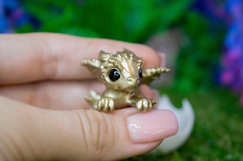 May include: A small, gold-colored dragon figurine with black eyes. The dragon is sitting on a hand, with its wings spread out.