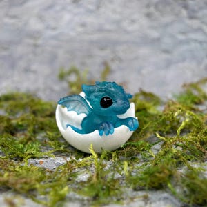 May include: A small, blue, translucent dragon figurine with black eyes, hatching from a white egg. The dragon is posed with its wings spread and its head tilted to the side. The egg is cracked open, revealing the dragon inside.