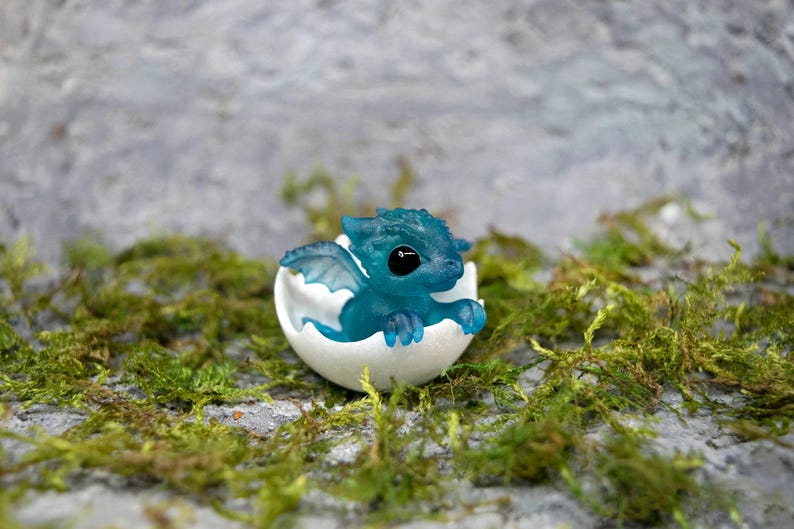 May include: A small, blue, translucent dragon figurine with black eyes, hatching from a white egg. The dragon is posed with its wings spread and its head tilted to the side. The egg is cracked open, revealing the dragon inside.