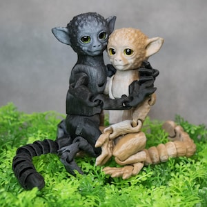 Könnte beinhalten: Zwei kleine, artikulierte Spielzeug-Affen. Einer ist schwarz mit blauen Augen und der andere ist braun mit gelben Augen. Sie umarmen sich und sitzen auf einem Bett aus grünem Kunstgras.