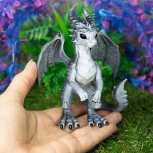 May include: A detailed figurine of a dragon with gray scales, wings, and horns. The dragon is perched on a hand, showcasing its intricate design. The background features artificial plants with purple and blue hues, creating a fantasy theme.