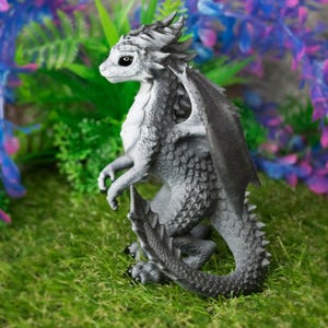 May include: A detailed figurine of a dragon in shades of gray, with textured scales and wings. The dragon is posed on a bed of green moss, with colorful artificial plants in the background. The dragon has a detailed face and is standing upright.