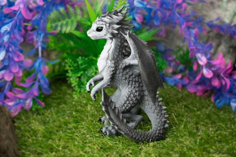 May include: A detailed figurine of a dragon in shades of gray, with textured scales and wings. The dragon is posed on a bed of green moss, with colorful artificial plants in the background. The dragon has a detailed face and is standing upright.