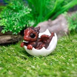 May include: A small, red dragon figurine hatching from a white eggshell. The dragon has black eyes and a small, curled tail. The figurine is sitting on a bed of green grass.
