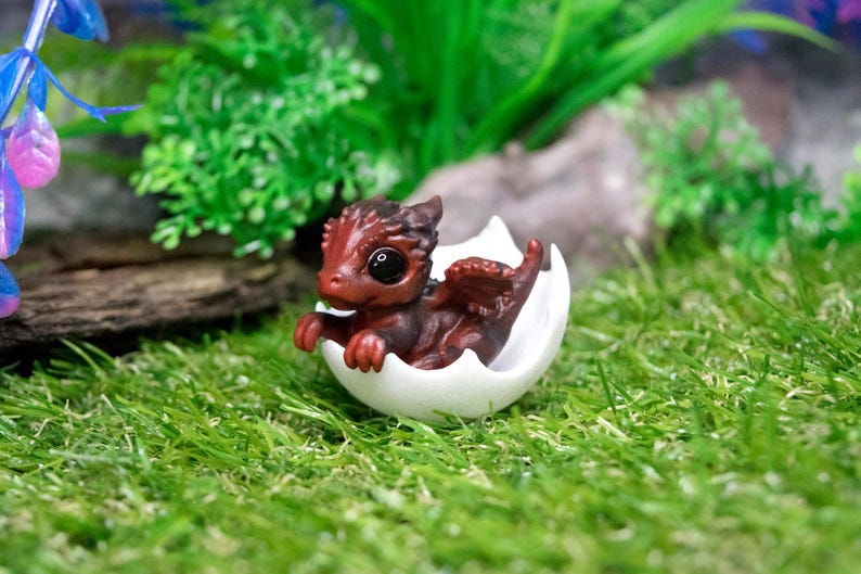 May include: A small, red dragon figurine hatching from a white eggshell. The dragon has black eyes and a small, curled tail. The figurine is sitting on a bed of green grass.