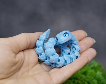 May include: A light blue, segmented, articulated snake toy with a blue eye. The snake is curled up in a hand.