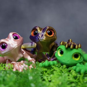 以下が含まれることがあります： 大きな目をした、カラフルな手描きの小さなカエルの置物3つ。カエルたちは緑の苔の上に座っています。