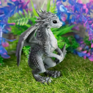 May include: A gray dragon figurine with detailed scales, wings, and a small tail, posed on a bed of green grass. The dragon has large, dark eyes and is surrounded by purple and blue artificial plants. The figurine is approximately 4 inches tall.