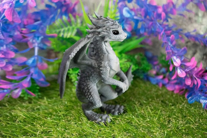 May include: A gray dragon figurine with detailed scales, wings, and a small tail, posed on a bed of green grass. The dragon has large, dark eyes and is surrounded by purple and blue artificial plants. The figurine is approximately 4 inches tall.