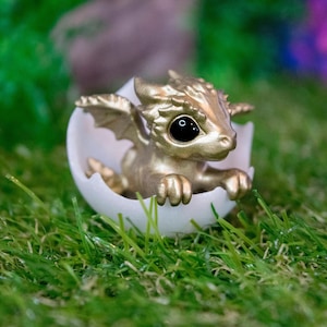 May include: A small, gold-colored dragon figurine is hatching from a white egg. The dragon has large, black eyes and is posed with its wings spread out. The figurine is sitting on a bed of green grass.