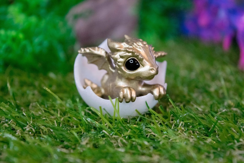May include: A small, gold-colored dragon figurine is hatching from a white egg. The dragon has large, black eyes and is posed with its wings spread out. The figurine is sitting on a bed of green grass.