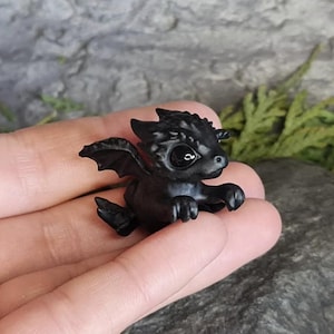 May include: A small black dragon figurine with a single black eye. The dragon has wings and is posed in a flying position.