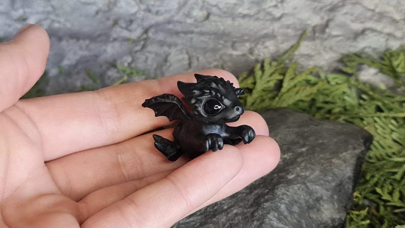 May include: A small black dragon figurine with a single black eye. The dragon has wings and is posed in a flying position.