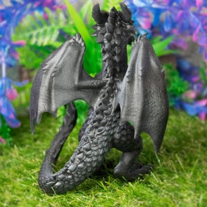 May include: A detailed, dark gray dragon figurine with outstretched wings, horns, and a textured body. The dragon is posed on a bed of green grass, with a backdrop of purple and blue artificial plants. The figurine is a fantasy-themed collectible.
