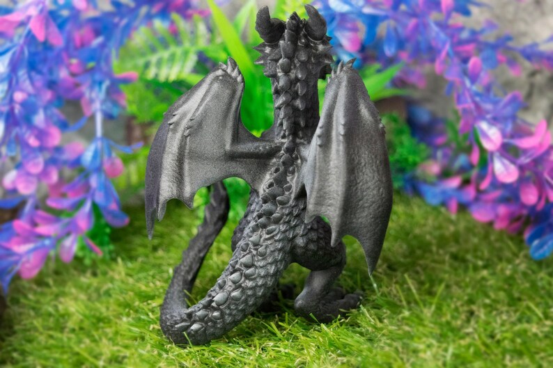 May include: A detailed, dark gray dragon figurine with outstretched wings, horns, and a textured body. The dragon is posed on a bed of green grass, with a backdrop of purple and blue artificial plants. The figurine is a fantasy-themed collectible.