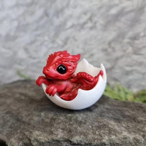 May include: A small, red dragon figurine hatching from a white egg. The dragon has black eyes and is posed in a curled position.