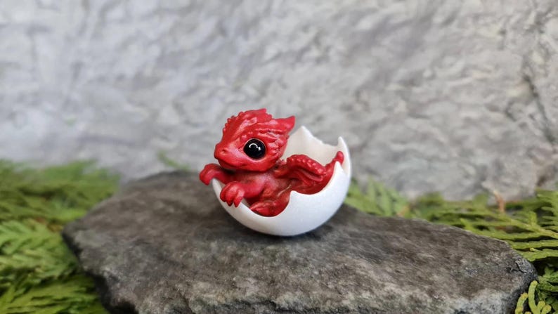 May include: A small, red dragon figurine hatching from a white egg. The dragon has black eyes and is posed in a curled position.