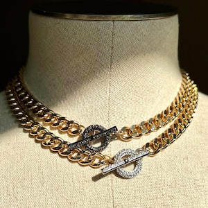 May include: Two gold-toned chain necklaces with toggle clasps. One clasp is adorned with black crystals, the other with clear crystals. The necklaces are displayed on a mannequin, showcasing their design and texture.