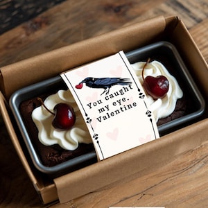 May include: A small brown box with a white card that says "You caught my eye. Valentine."  The card has a black raven with a red heart in its beak. The box contains a chocolate brownie topped with whipped cream and a cherry.