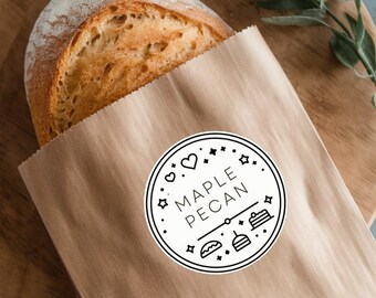 Könnte beinhalten: Eine braune Papiertüte mit einem weißen Aufkleber, auf dem in einem Kreis mit einem Herzen und anderen dekorativen Elementen "Maple Pecan" steht. Ein Brotlaib befindet sich teilweise in der Tüte.