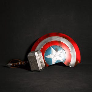 May include: A weathered Captain America shield with a broken edge, featuring red, white, and blue colors and a central white star. Next to it is a silver and gray Mjolnir hammer with a brown leather handle. The items are on a dark background.