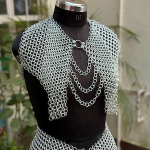 Puede incluir: Conjunto de top y falda de cota de malla plateada exhibido en un maniquí negro. La cota de malla está hecha de pequeños anillos metálicos entrelazados. El top presenta un cierre circular y un diseño de cadena drapeada. La falda es visible en la parte inferior del maniquí.