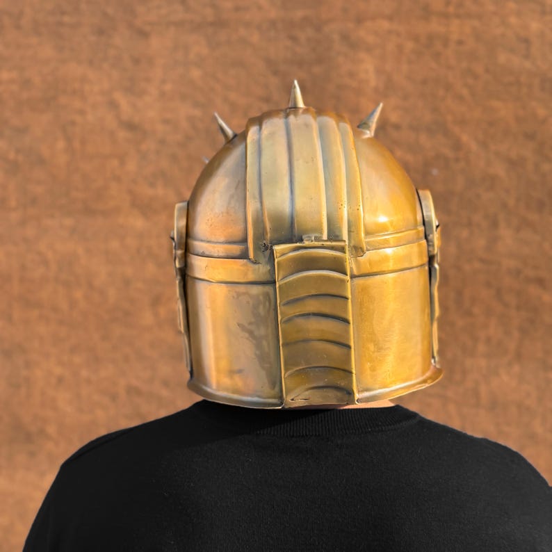 May include: A gold-colored helmet with a pointed top and spikes. The helmet has a textured surface and is worn by a person.
