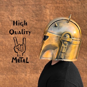 May include: A gold-colored metal helmet with a pointed top and a raised design on the side. The helmet is worn by a person with a black shirt. The text "High Quality Metal" is visible in the image.