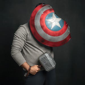 May include: A Captain America shield with a red, white, and blue design, and a silver hammer with a brown handle. The shield has a white star in the centre. The items are held by a person wearing a grey sweatshirt.