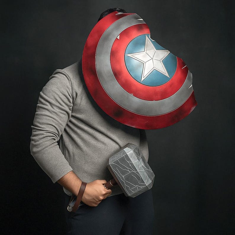 May include: A Captain America shield with a red, white, and blue design, and a silver hammer with a brown handle. The shield has a white star in the center. The items are held by a person wearing a gray sweatshirt.