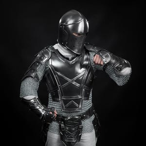 May include: A complete set of medieval armour, featuring a helmet, chest plate, and arm guards. The armour is a dark, metallic hue with detailed patterns and chainmail sections. The helmet has a visor, and the armour covers the entire body.