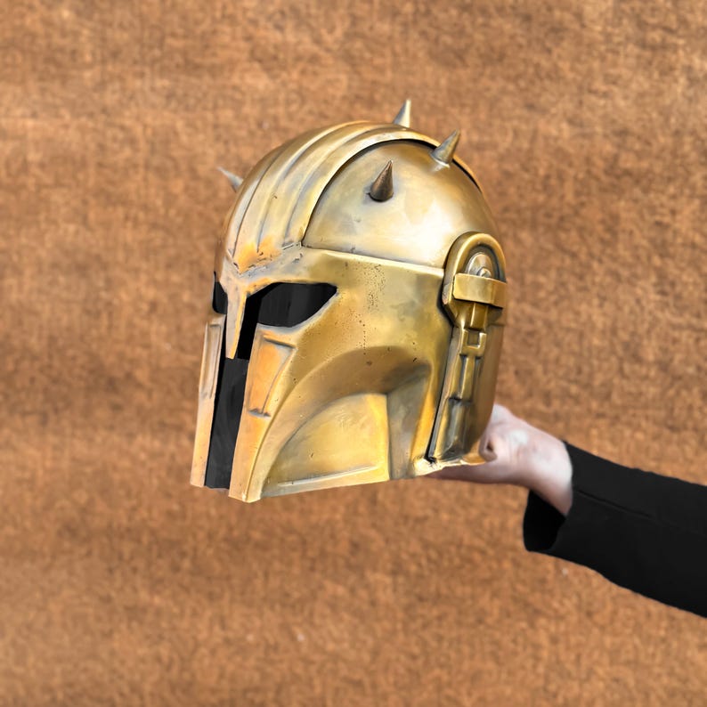 May include: A gold-colored helmet with a black visor and spikes on top. The helmet is made of metal and has a textured surface.