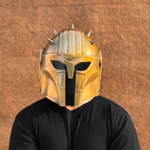 May include: A gold-colored helmet with a pointed design and spikes. The helmet has a dark visor and is worn by a person with a black shirt.