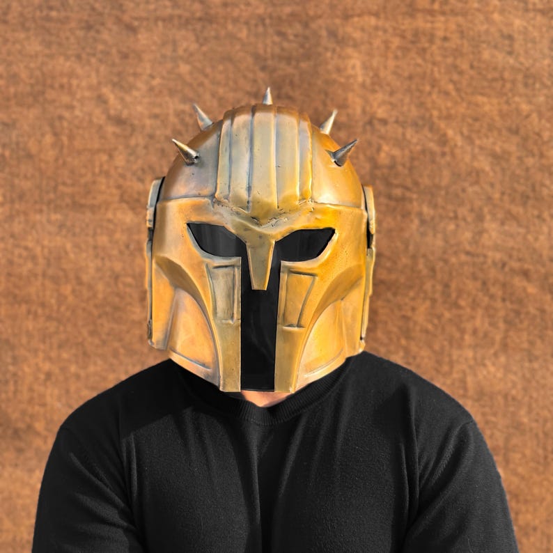 May include: A gold-colored helmet with a pointed design and spikes. The helmet has a dark visor and is worn by a person with a black shirt.