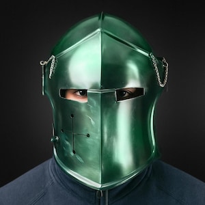 Puede incluir: Un casco medieval verde metálico con un diseño de cruz en la parte delantera. El casco tiene ranuras para los ojos y un detalle de cadena en el lateral. La persona lleva una sudadera azul oscuro.