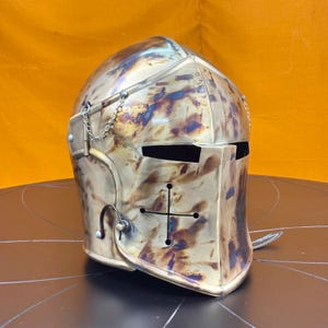 May include: A medieval helmet with a cross design on the front, made of metal with a distressed finish. The helmet has a chainmail chin strap and a visor.