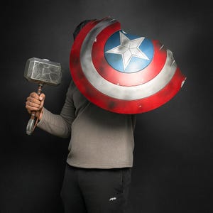 May include: A Captain America shield with a five-pointed star in a blue circle, surrounded by red and white stripes. A silver hammer with a wooden handle is held in the other hand. The shield has a distressed, battle-worn appearance.