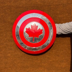 May include: A red, white and silver shield with a red maple leaf in the centre. The shield is round and has a raised edge.