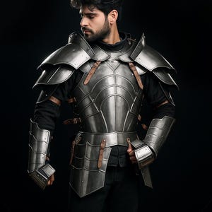 Könnte beinhalten: Eine silberfarbene mittelalterliche Rüstung mit Lederriemen. Die Rüstung umfasst eine Brustplatte, Schulterpanzer und Armschützer. Die Person auf dem Bild trägt ein schwarzes Hemd und eine schwarze Hose. Der Hintergrund ist schwarz.