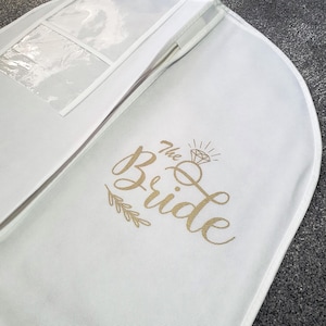 May include: White garment bag with a clear plastic window. The bag has the words "The Bride" embroidered in gold with a diamond graphic.