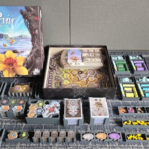 May include: A board game called "Ränne" with a box lid featuring a fantasy landscape and a flower. The game board is laid out with various components, including cards, tokens, and dice. The game is organised in a variety of plastic storage containers.