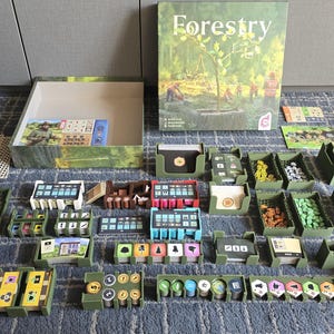 Może przedstawiać: Gra planszowa Forestry zawiera duże ilustrowane pudełko, puste pudełko na grę oraz liczne elementy. W skład zestawu wchodzą zielone plastikowe podstawki na karty, stosy małych okrągłych żetonów w różnych kolorach, prostokątne karty oraz płytki z siatką.