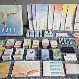 May include: A board game called "KRILE" with a blue, green, and white game board, colorful playing cards, and a box with a bird illustration. The game includes a variety of components, including wooden pieces, dice, and cards.