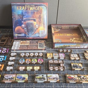 Puede incluir: Juego de mesa "Kraftwagen" con componentes variados: cartas, fichas y tablero. La caja muestra un coche antiguo y un hombre. Las piezas del juego están dispuestas sobre una superficie estampada.