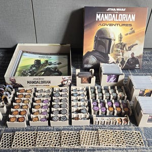 Op de afbeelding: Star Wars The Mandalorian Adventures bordspelset inclusief een grote speldoos, een spiraalgebonden regelboek, meerdere beige plastic organizers, stapels spelkaarten en talloze ronde fiches met diverse symbolen en kleuren.
